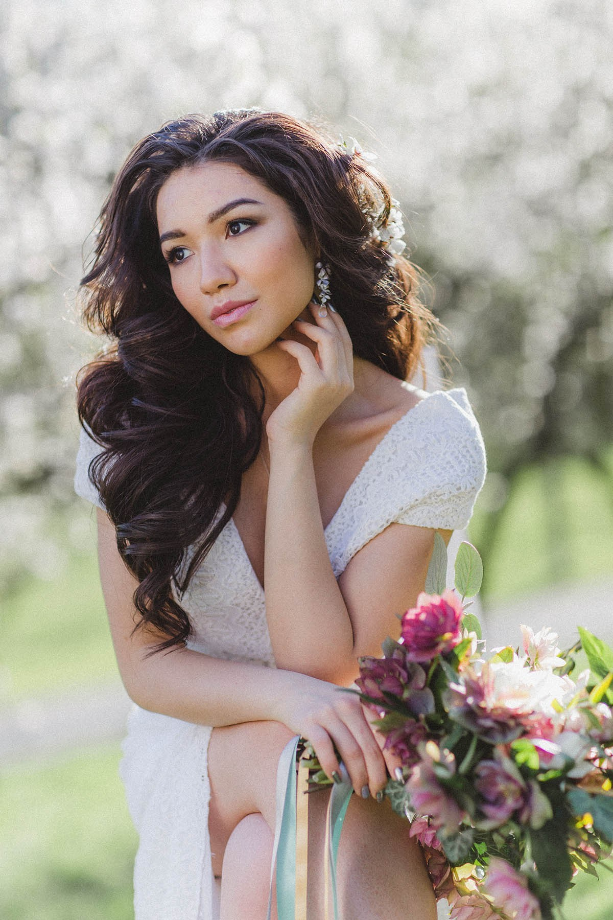 Beautiful young brunette, sitting crossed-legged, holding a bouquet, looking away under Cherry Blossom Trees, Prague.