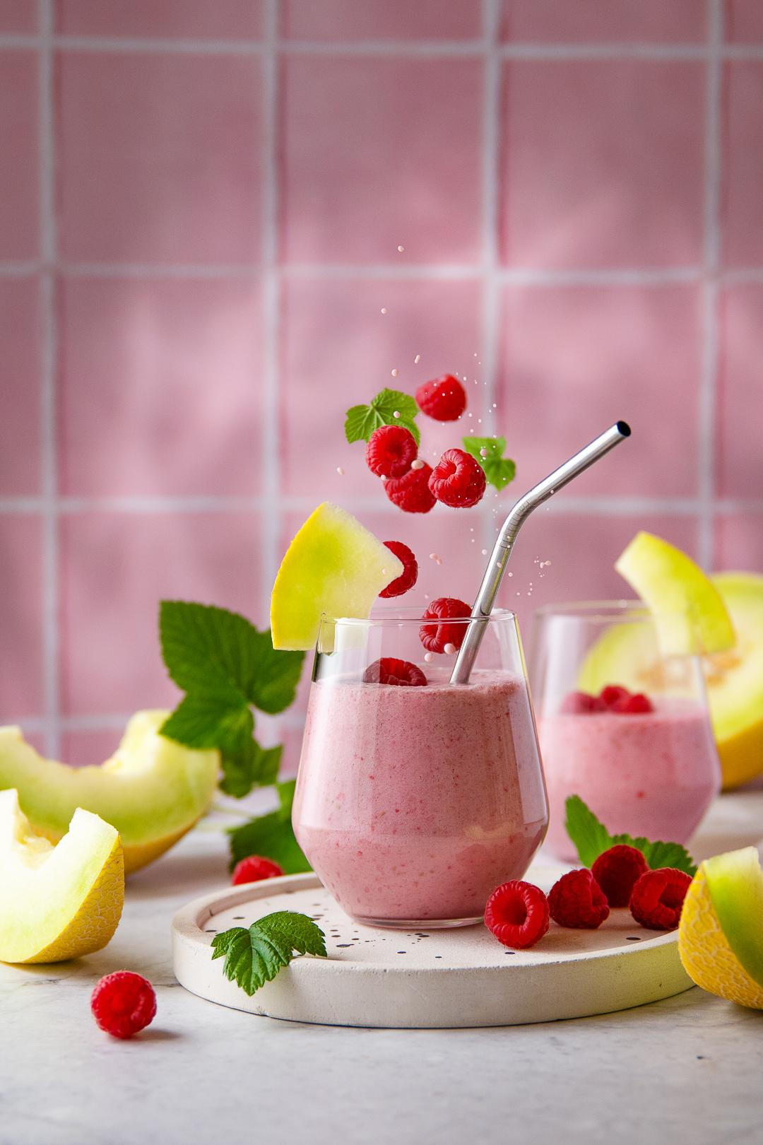 Ein sommerliches Getränk in der Kombination, Himbeeren, Melone, Kokosmilch und Ingwer. Das Getränk hat eine cremige Textur und einen reichen Beerengeschmack, serviert in zwei Gläsern mit vielen Himbeeren und frischer Melone vor einem rosa Hintergrund