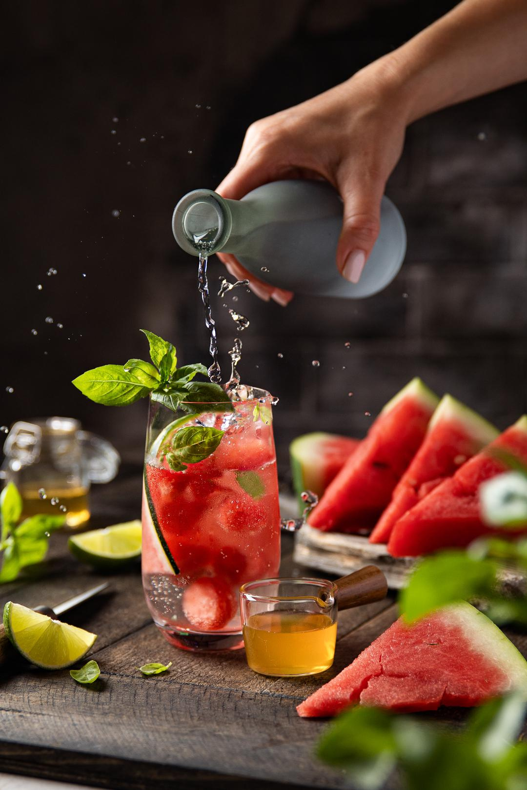 Wassermelonen-Mojito im Glas mit Basilikum, Limette, Honig und ausgegossenes Mineralwasser aus der Flasche auf dem Holzbrett