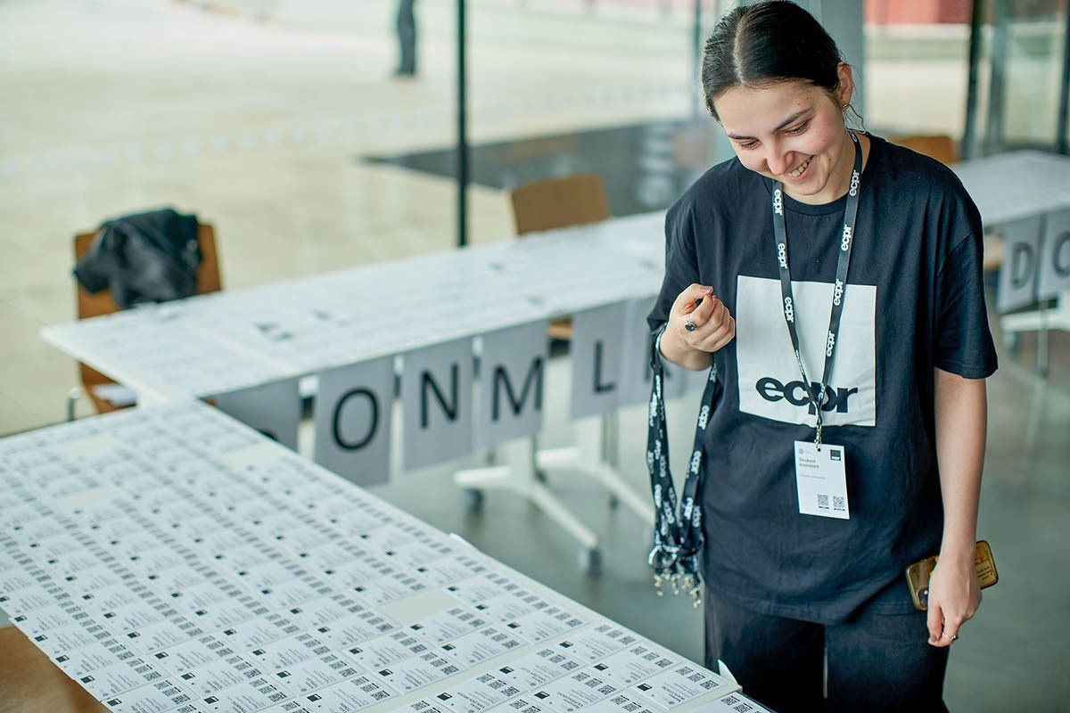 A smiling woman holding lanyards organizes a table of conference names before the educational event kickoff in Prague.