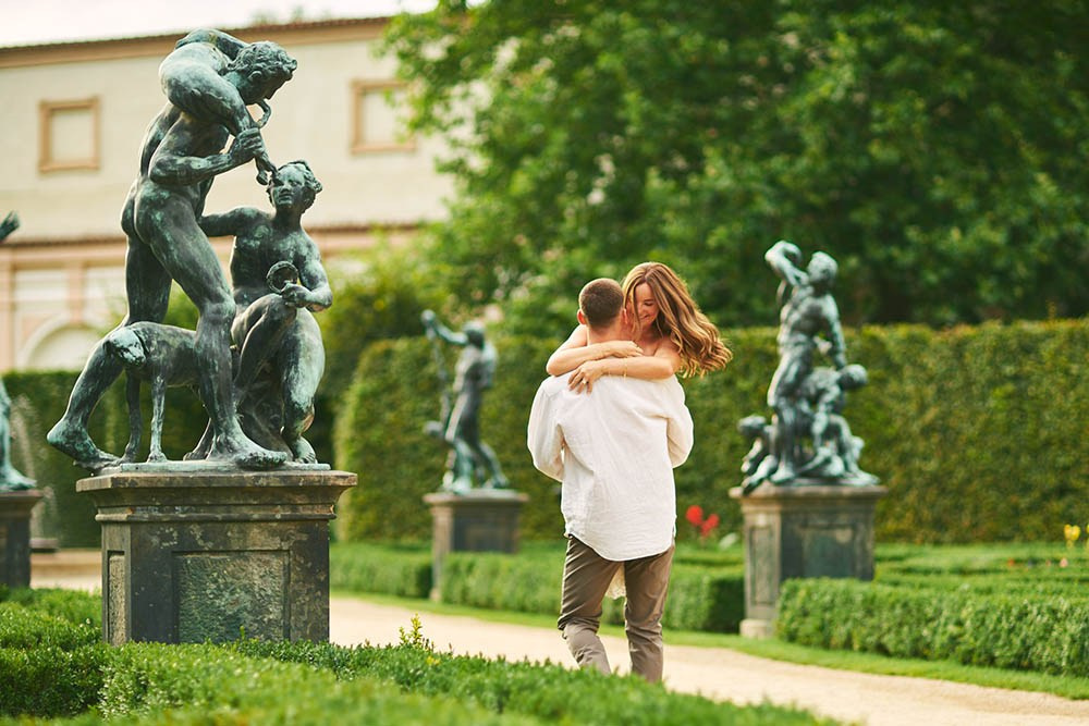 A woman is carried aloft by her fiancé near the historic statues in the Wallenstein Garden, Prague