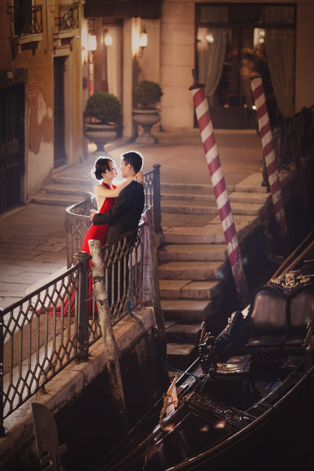 A Thai woman wearing a red dress and caressed by her fiancee are nearly silhouetted in a night time portrait captured near a gondola in Venice.