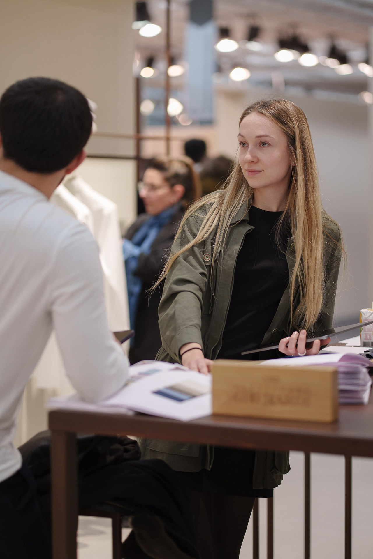 A fashion consultant at a desk assisting a visitor, surrounded by luxury garments and lookbooks.
