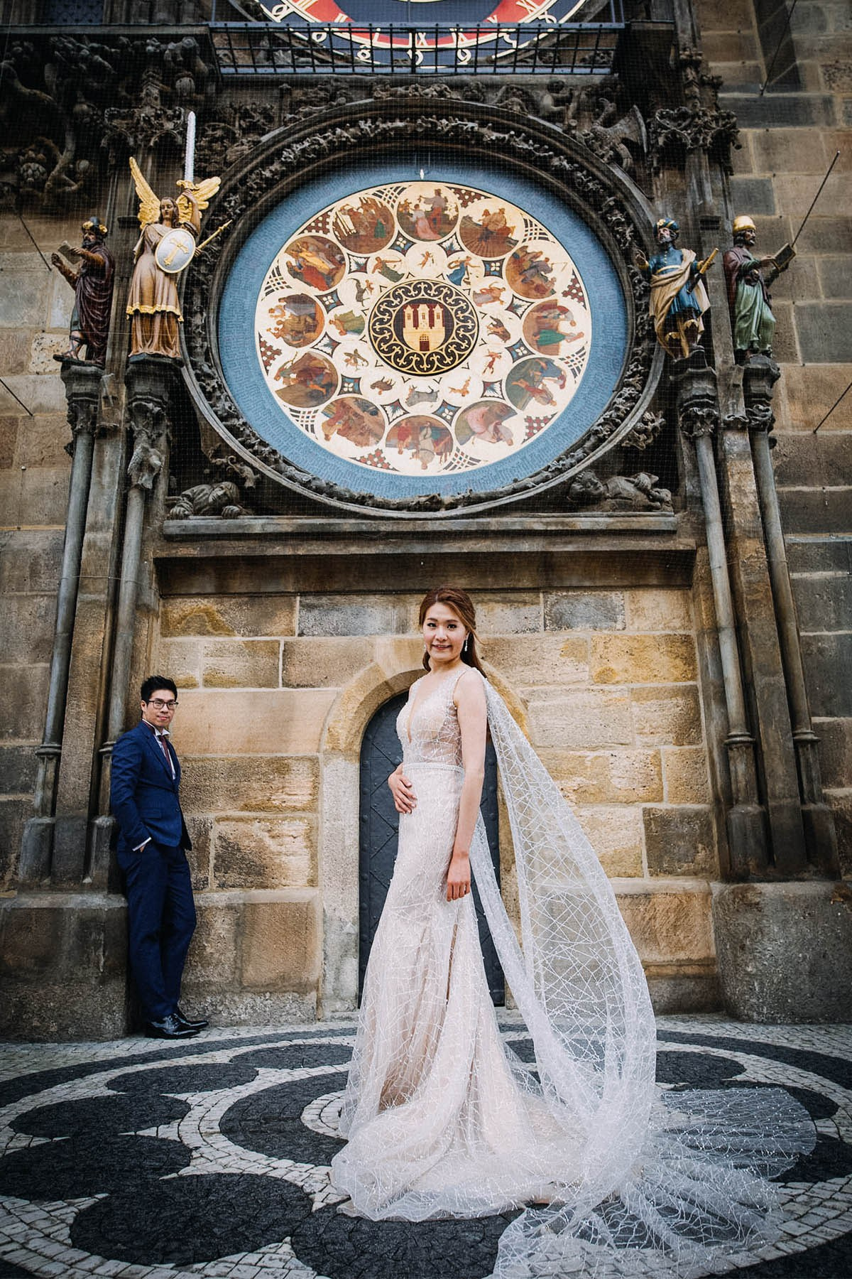 Woman in elegant evening wear stands in pose underneath Prague's Astronomical Clock as her partner looks on from behind.