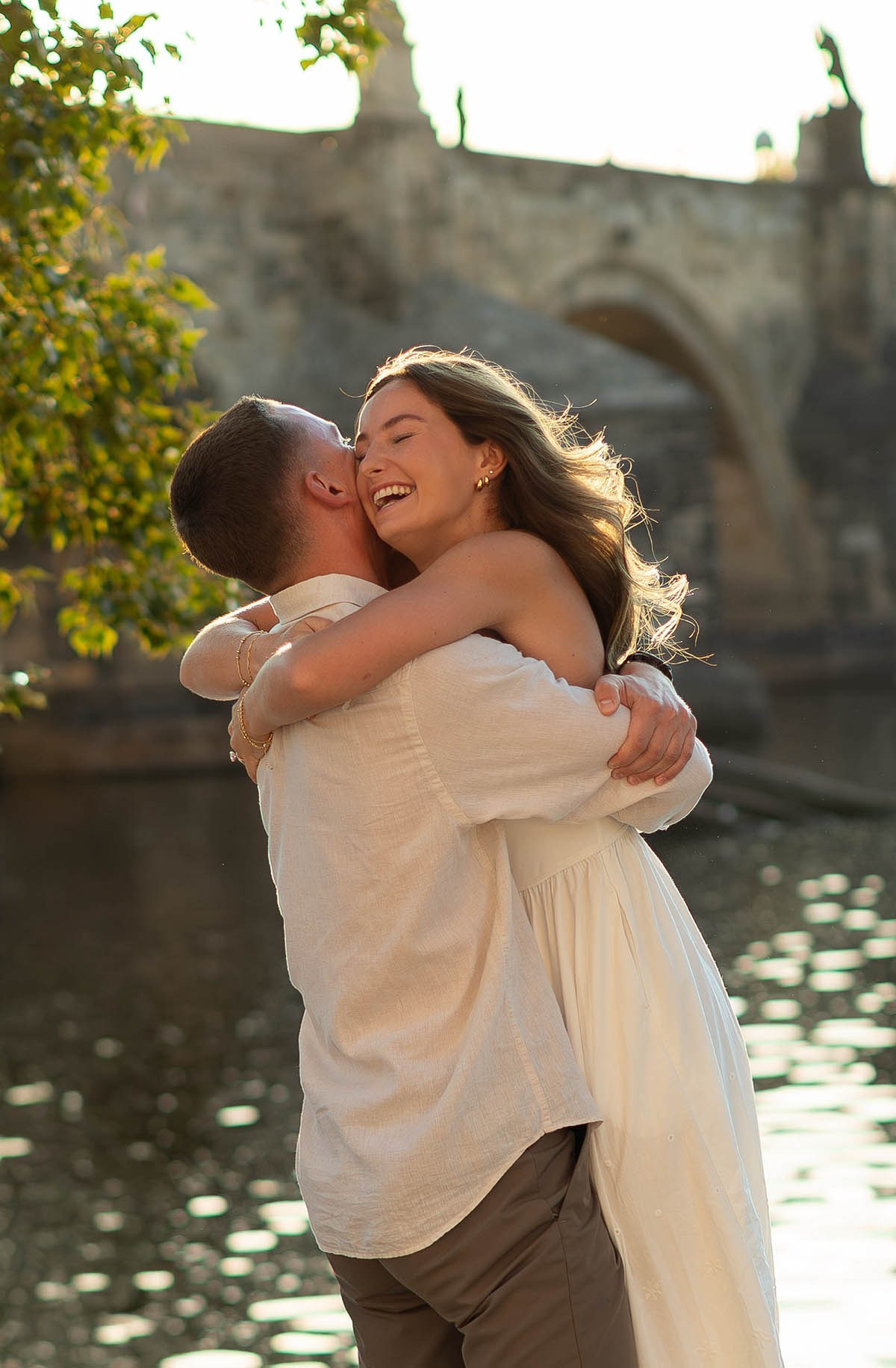 A smiling, happy woman jumps into her partner's arms after he proposed to her near the Charles Bridge.