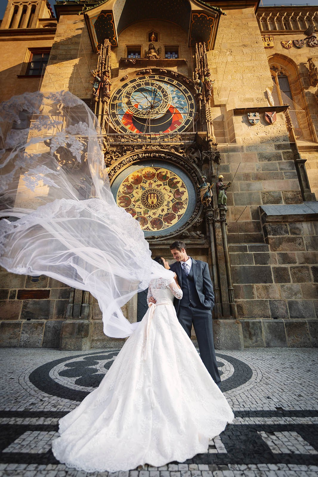 A bride and groom stand underneath the Astronomical Clock in Prague as her veil floats high in the air