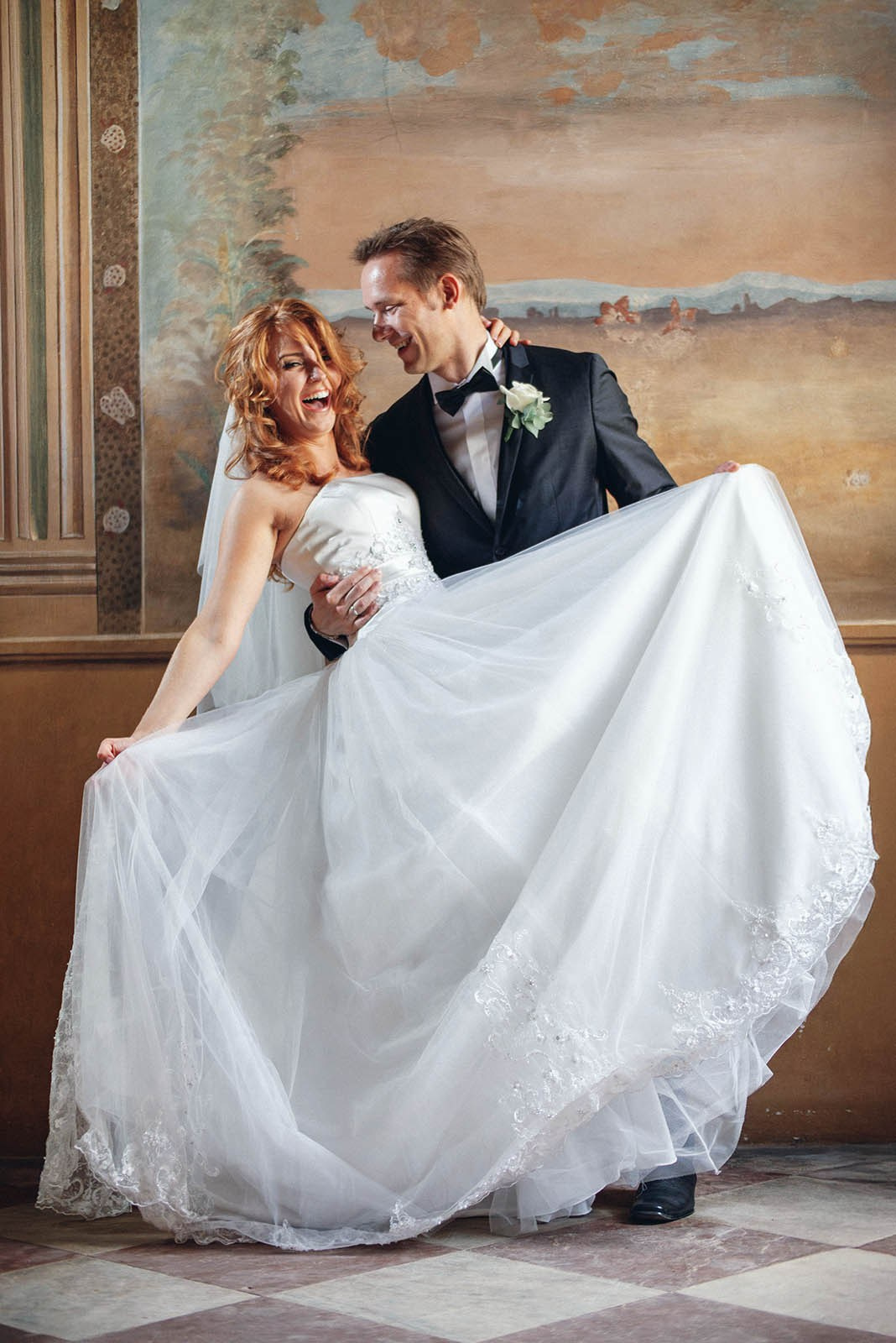 Red-headed bride laughing out loud as her groom dips her while holding her wedding dress at the Vrtba garden in Prague