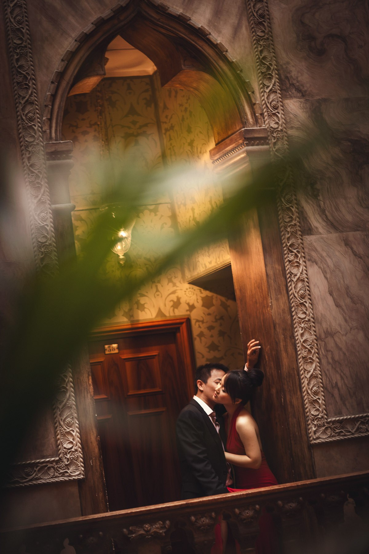 A woman wearing a red dress is kissed by her Thai fiancee inside the historic interior of the Hotel Danieli in Venice during their Love Story portrait session.