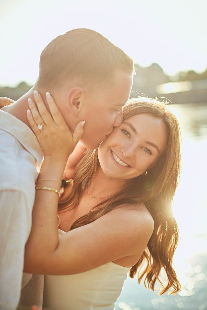 A smiling woman looks into the camera as her partner kisses her cheek as they are surrounded by sun flare near the riverside.