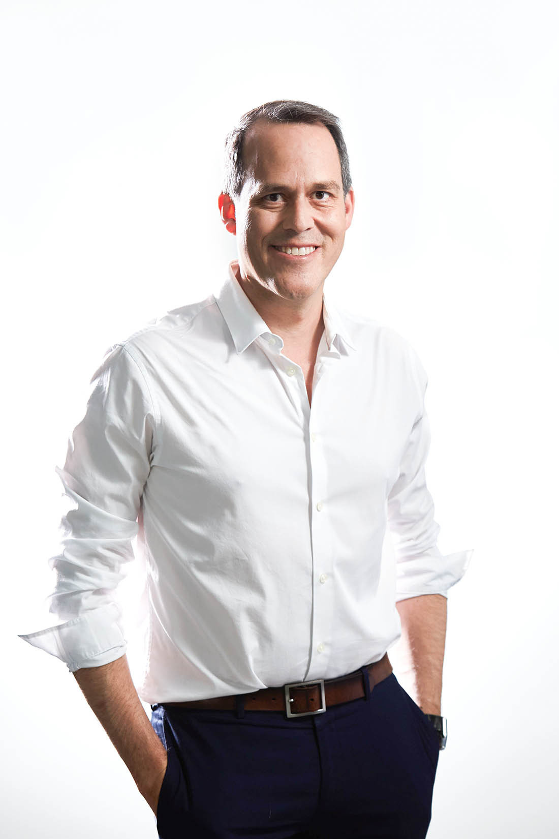 A smiling man wearing a white collared buttoned shirt smiles in a relaxed pose against a white background.