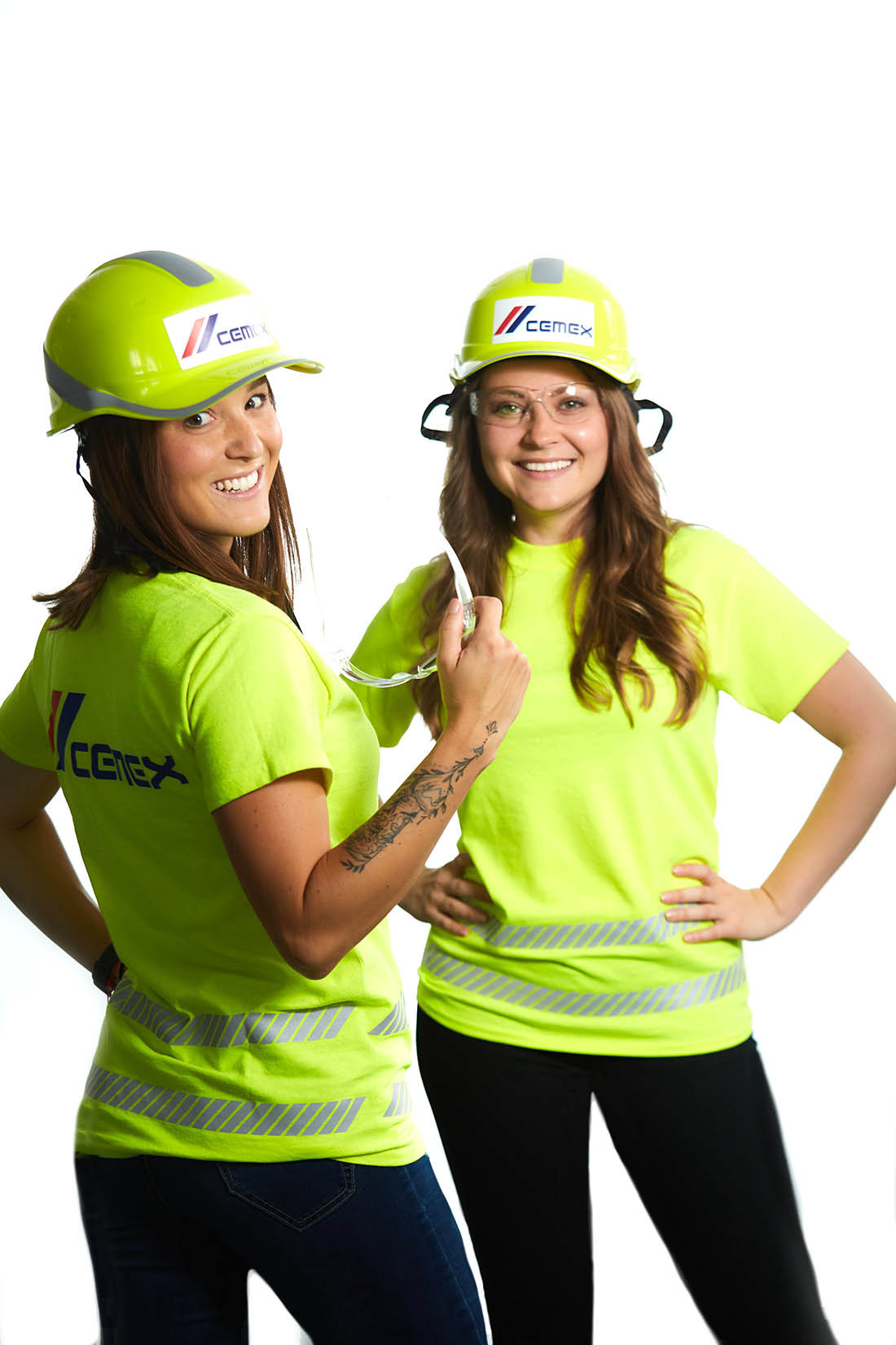 A portrait of two woman wearing safety glasses, yellow reflective jerseys and hard hats taken against a white studio wall. The portrait will be used for internal and external marketing and PR uses.