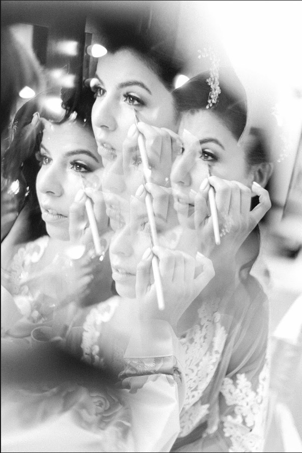 Multiple reflections of the bride's face in mirrors while applying makeup with a brush.