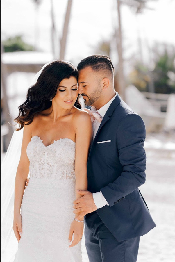The bride and groom stand with their heads bowed to each other, against the background of a blurry landscape with palm trees.