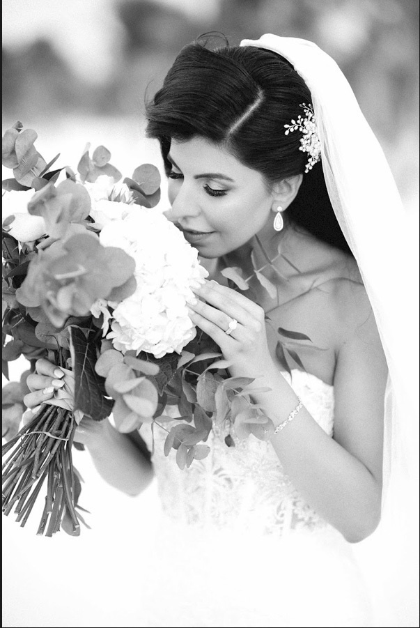The bride inhales the fragrance of white flowers of a large bouquet, which she holds in her hands. A white barrette stands out on her dark hair.