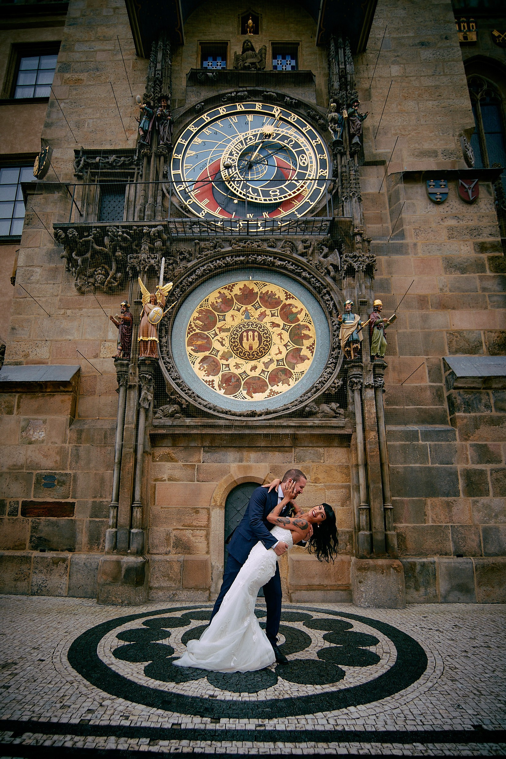 Groom dipping tattooed bride under historic Prague Astronomical Clock.
