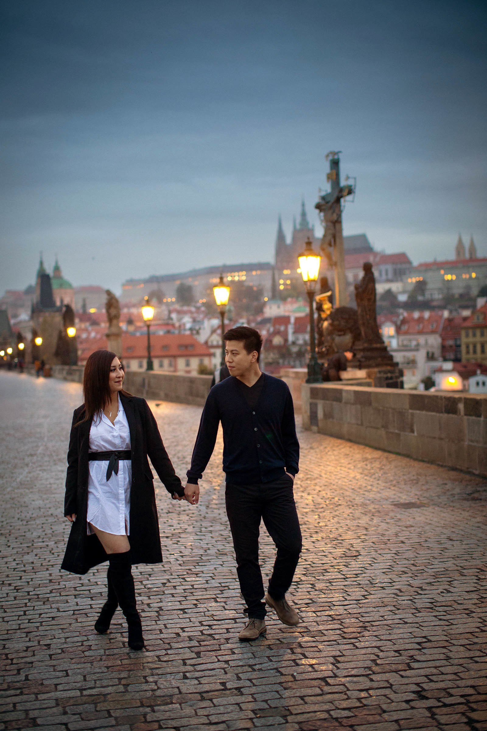 Smartly dress, dark-haired couple walk along the cobblestones atop the gas-lamp lit Charles Bridge at dawn