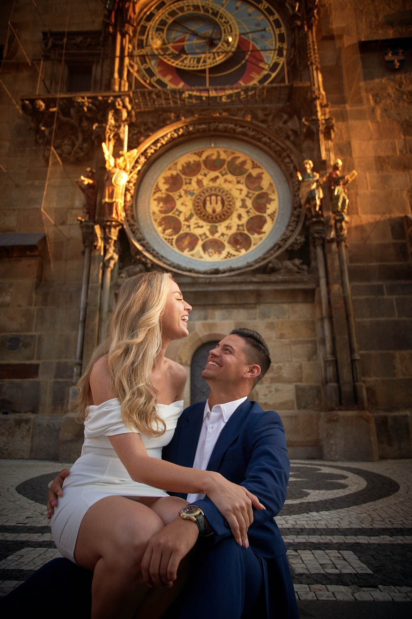 Gorgeous woman sitting in her partner's lap, laughing as they sit underneath the Astronomical Clock in Prague.