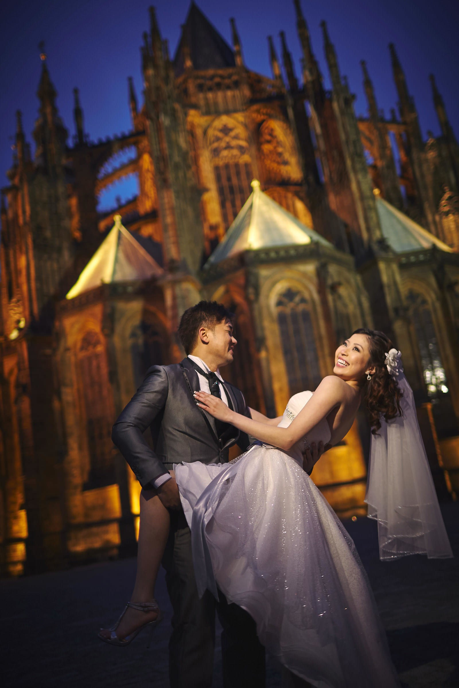 Vivacious Asian bride smiles as she is dipped by her stylish groom behind St. Vitus Cathedral at dusk