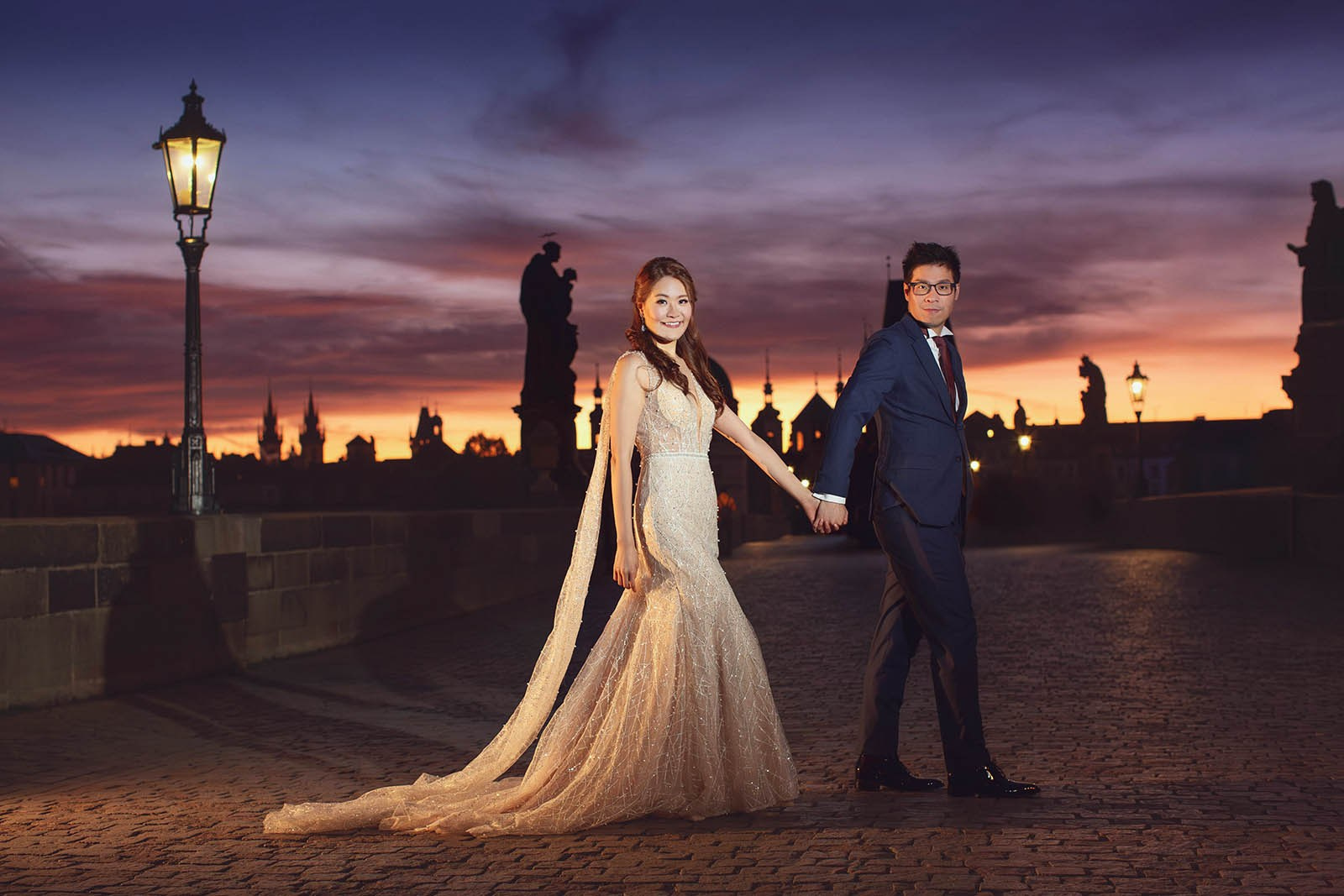 American couple held hands atop Charles Bridge at dawn with colorful sky, Prague