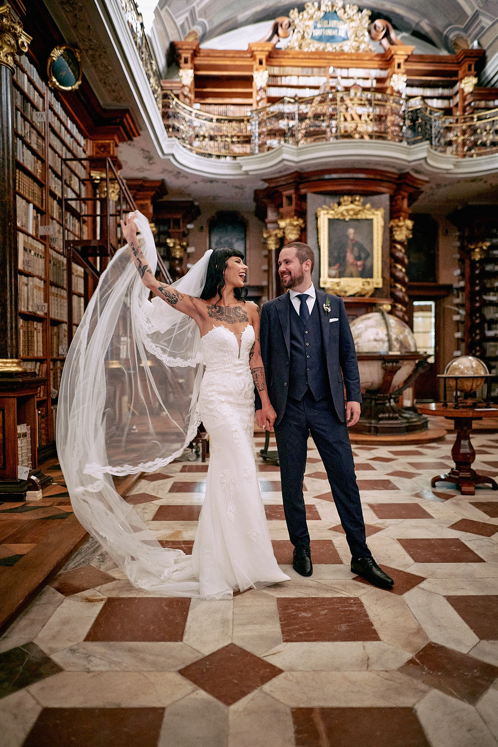 Tattooed bride gracefully lifting veil amid Klementinum Library shelves.