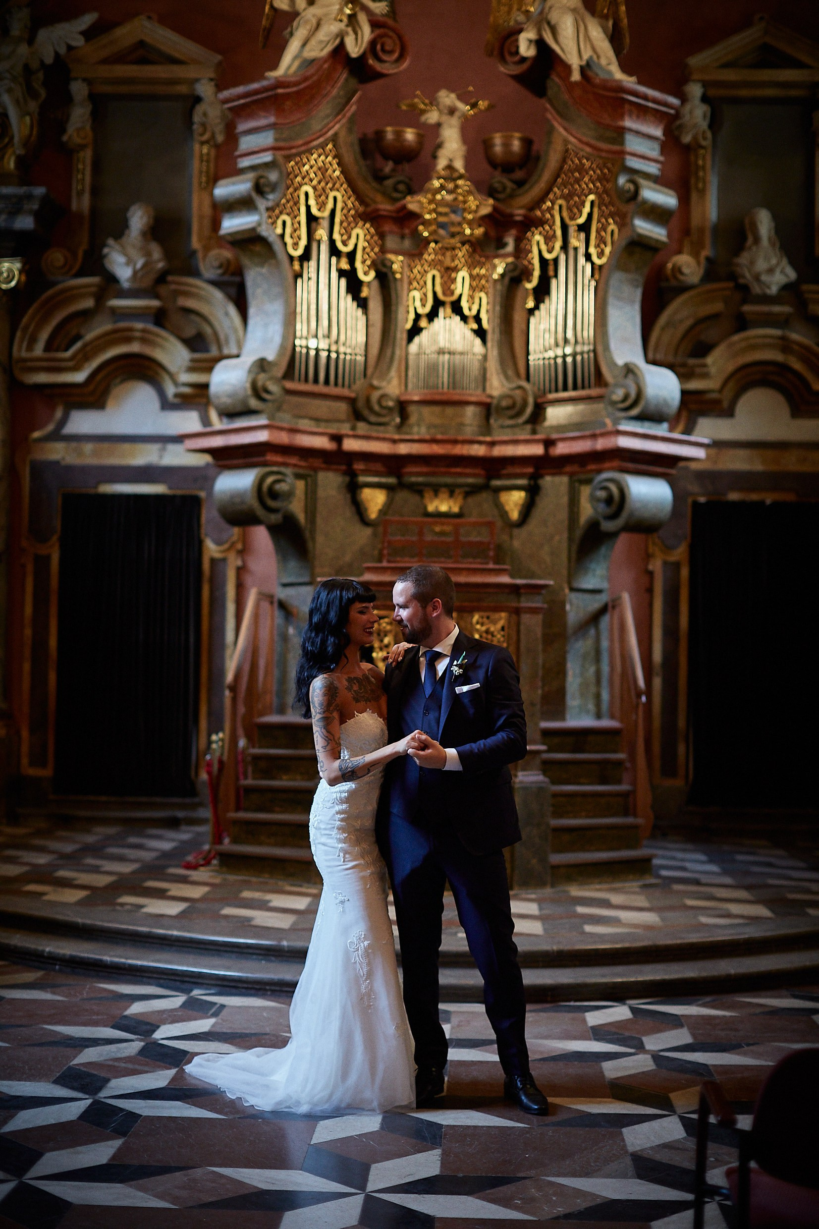 Groom hugging tattooed bride tenderly in baroque chapel setting.