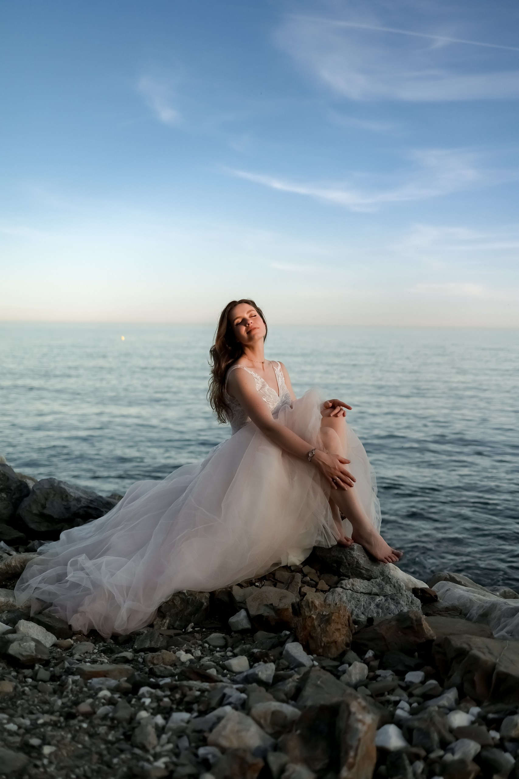 Braut sitzt solo auf Felsen am Mittelmeer, Brautkleid fließt, blauer Himmel, After Wedding Shooting Costa Blanca