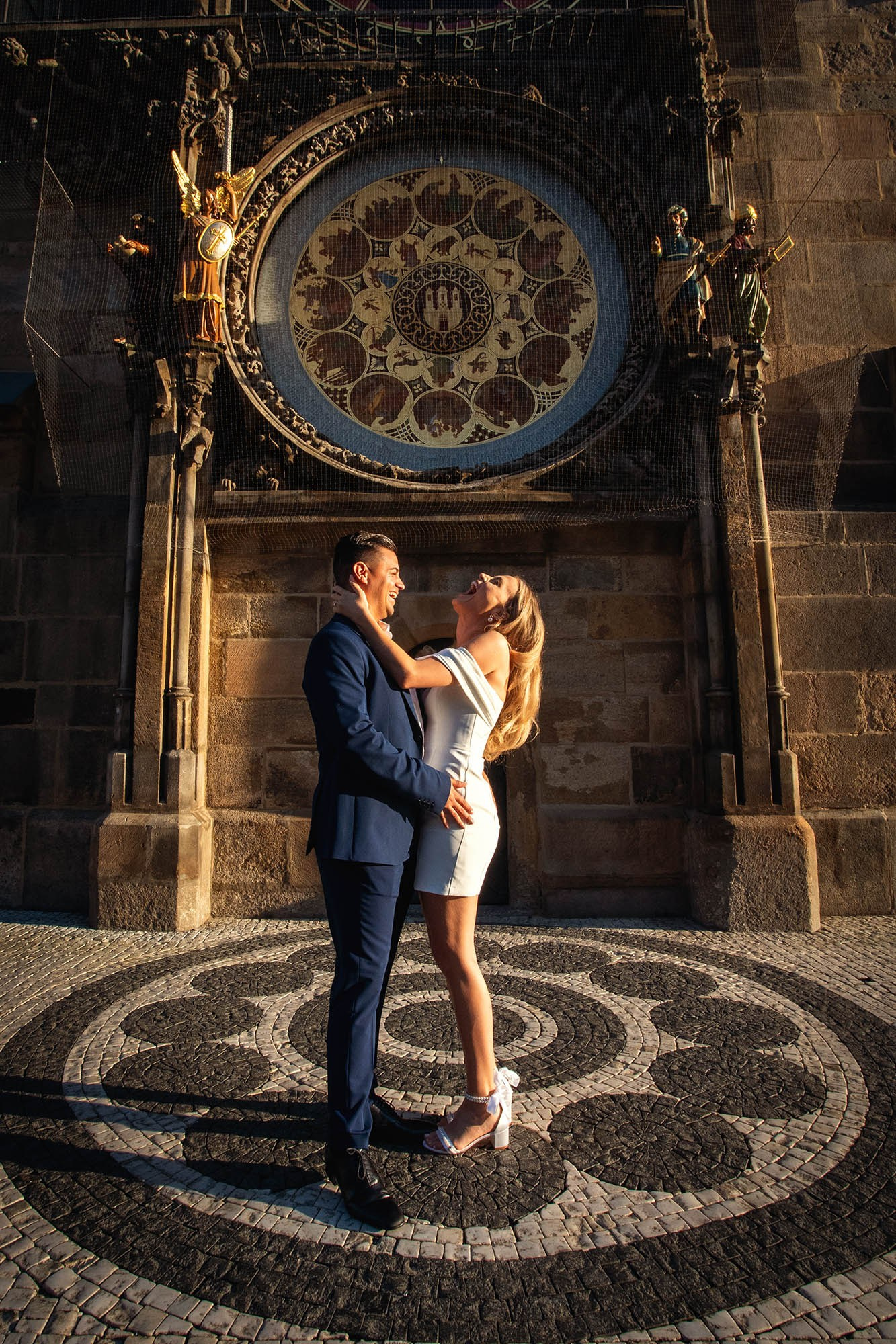 Blonde woman laughing joyfully in fiancé's arms under the Astronomical Clock at sunrise.