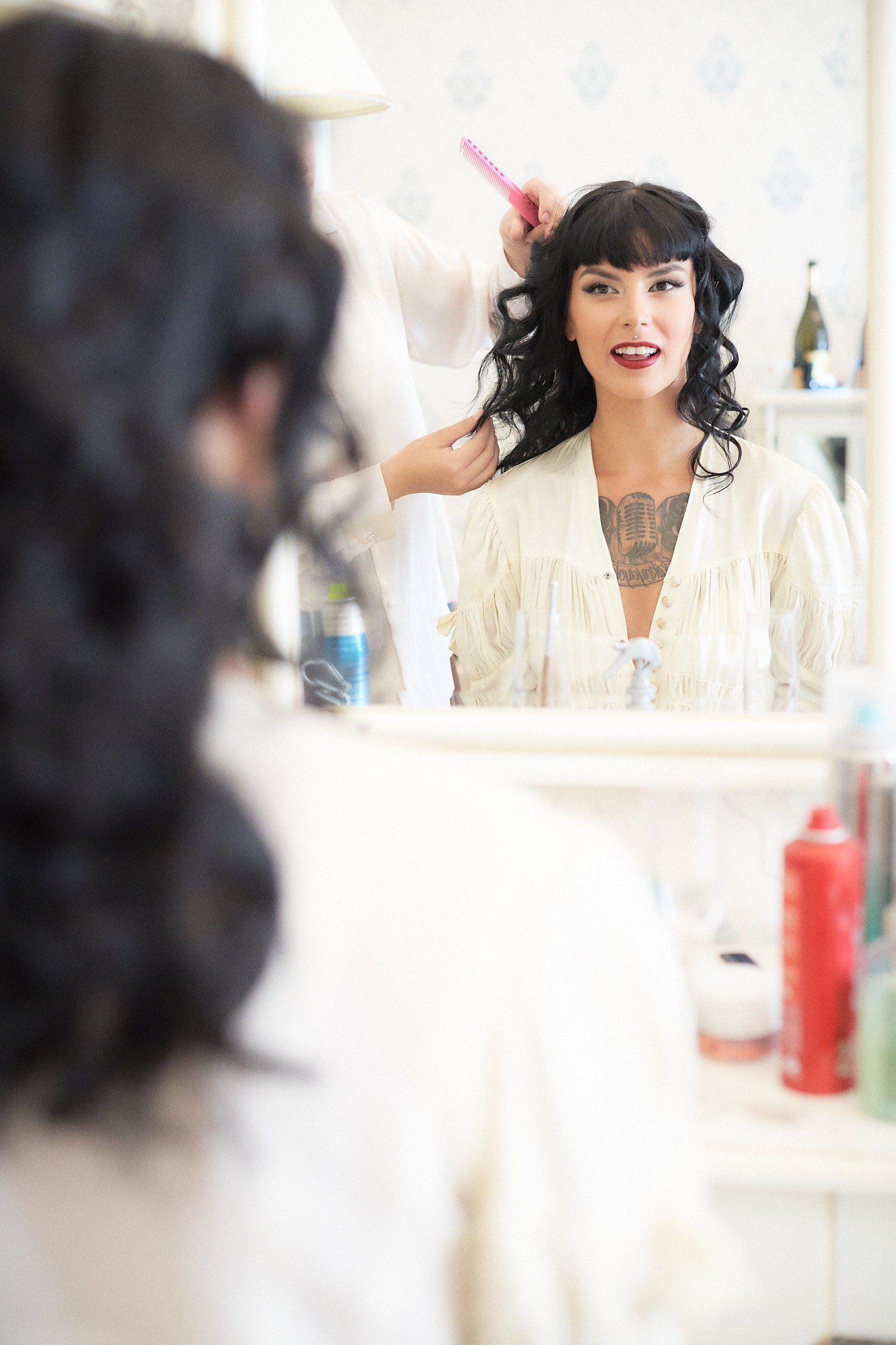 Tattooed bride observing hair and makeup finale in elegant suite.