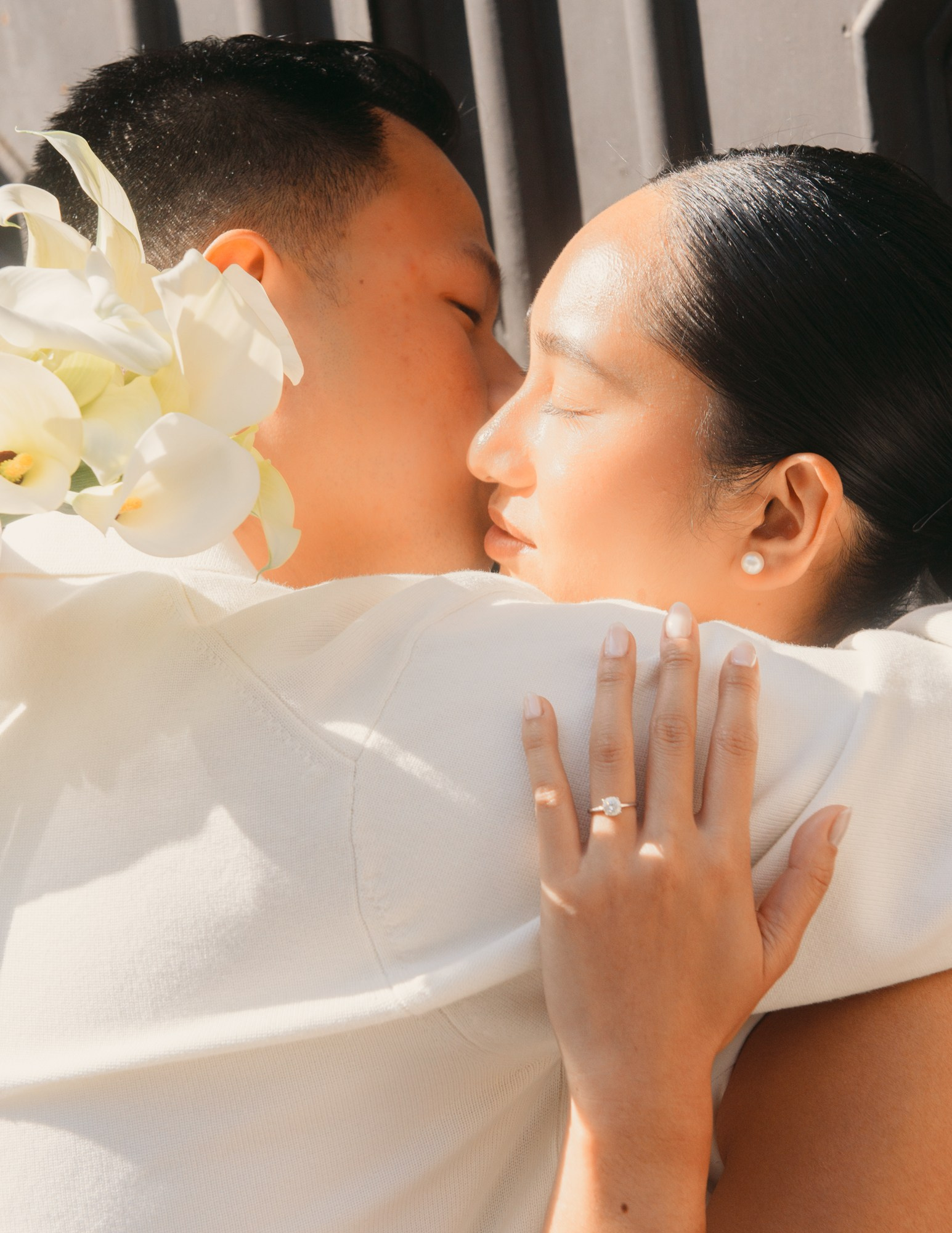 Romantic Engagement Portrait in Natural Light – Elegant Couple Photography. Close-up of an engaged couple embracing in soft natural light, with the bride showing her engagement ring and holding white flowers. A tender engagement portrait of a stylish couple wrapped in soft daylight, highlighting intimacy, love, and elegance. A timeless pre-wedding photography moment filled with emotion and natural beauty. engagement photography, romantic couple portrait, pre wedding photoshootnatural light photography, love story photography, intimate couple moment,engagement ring detailwedding photography inspiration, modern engagement session, elegant couple portrait
