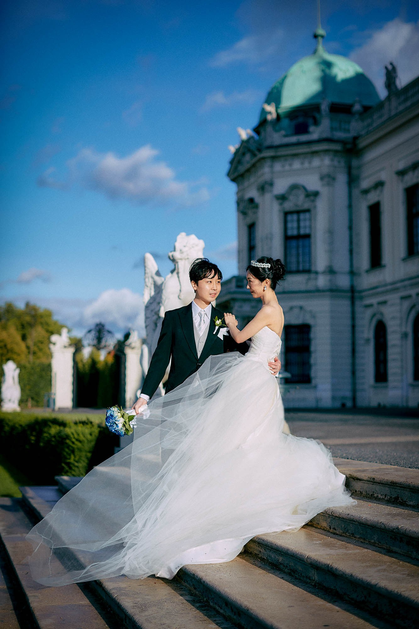 Newlyweds in sunlight near statue atop Belvedere Palace steps.