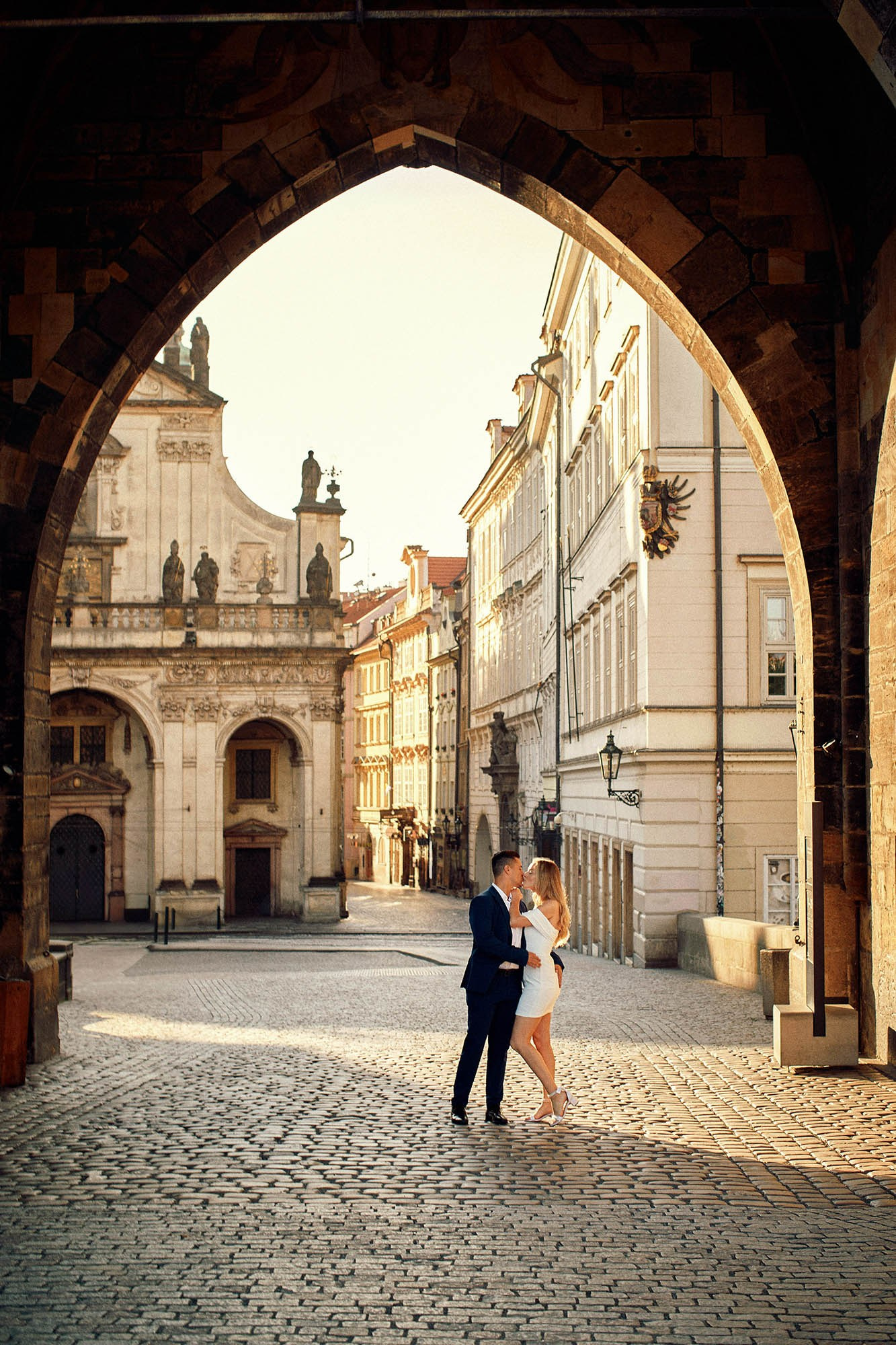 A couple sharing a soft kiss under a Gothic tower near Charles Bridge in early sunlight.
