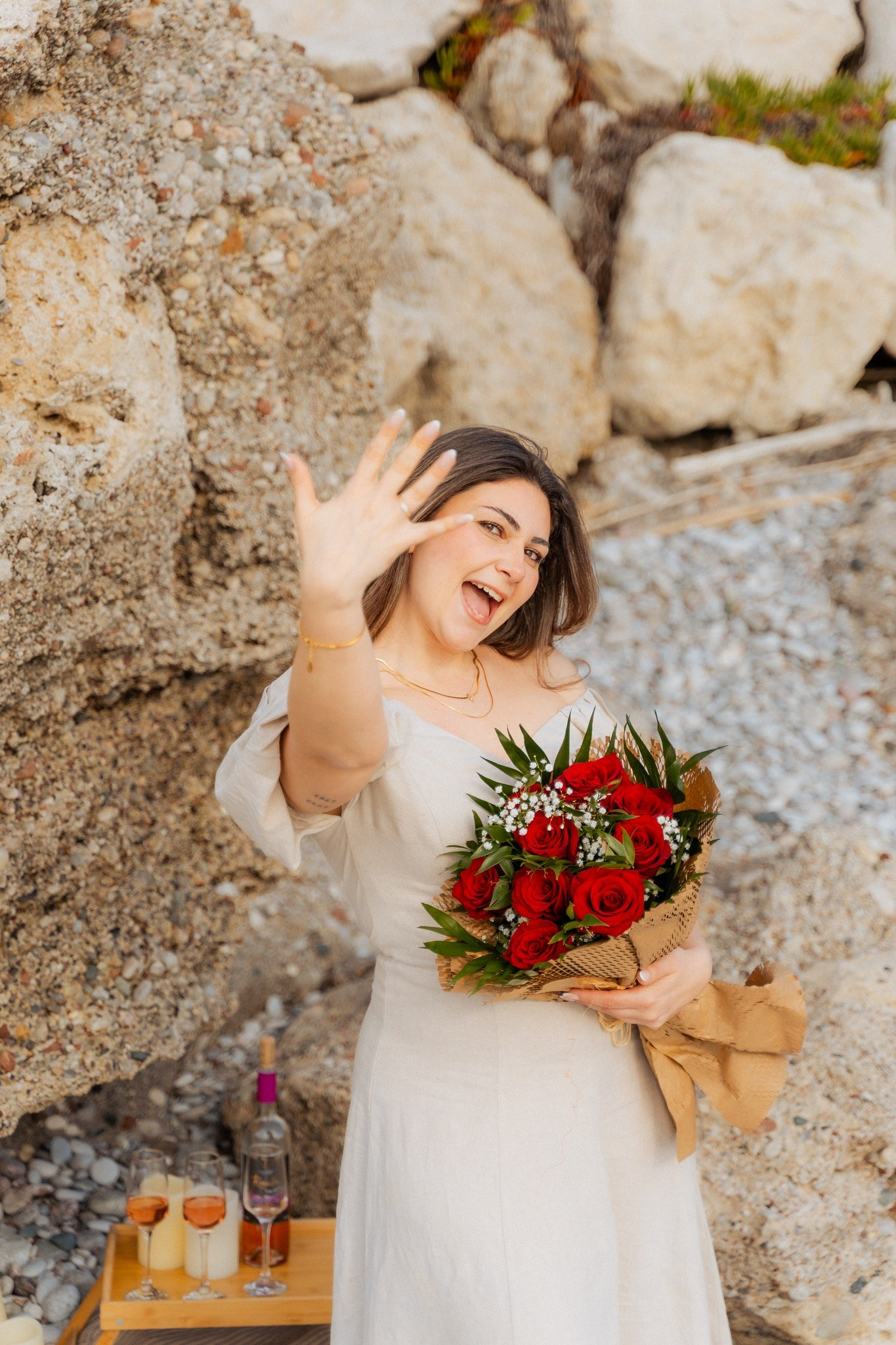 Junge Frau in hellem Kleid zeigt stolz ihren Verlobungsring am Strand von Rhodos und hält einen Strauß roter Rosen vor einer Felskulisse.
