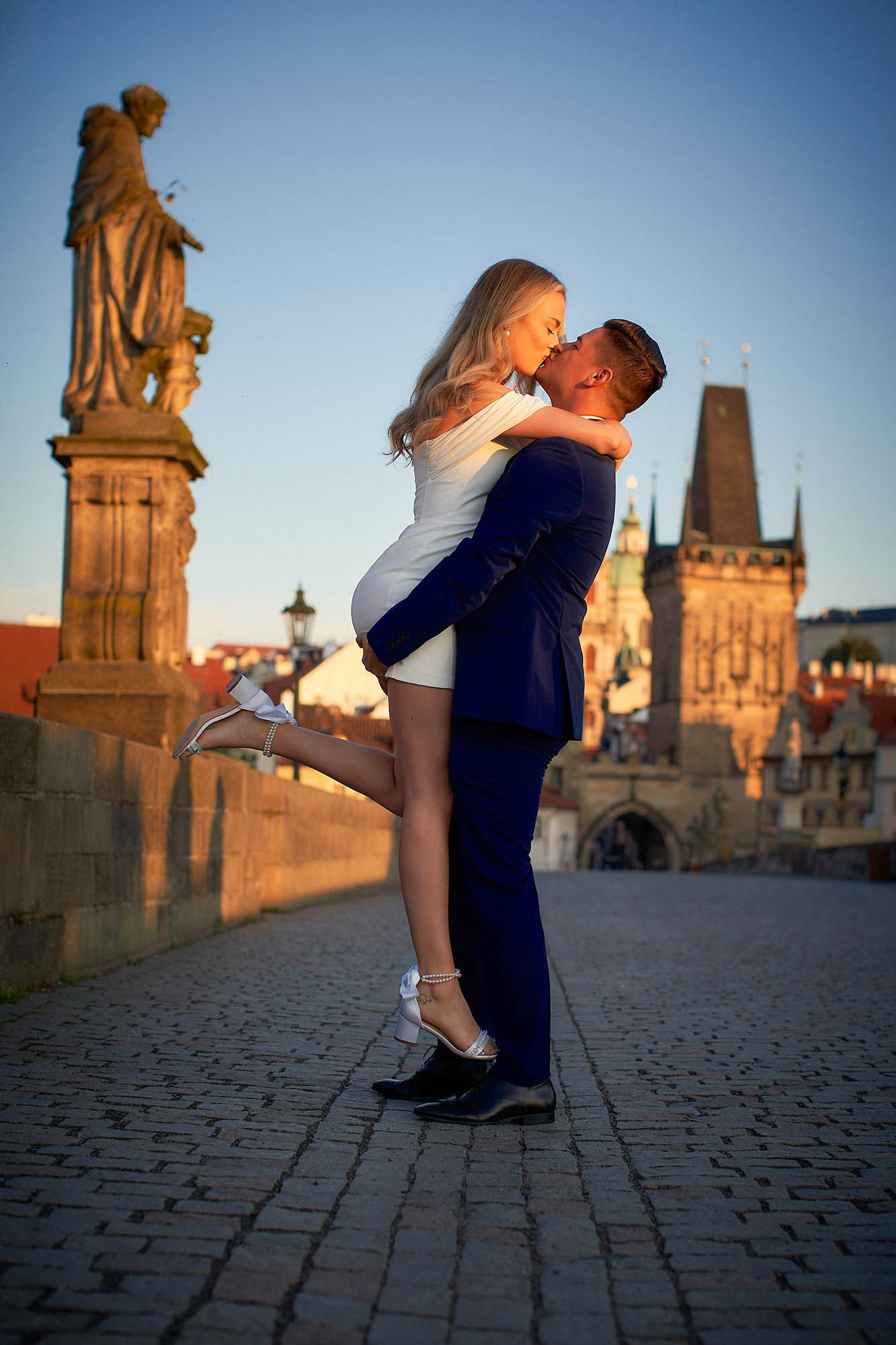 Groom lifting blonde fiancée for a passionate sunrise kiss on Charles Bridge.