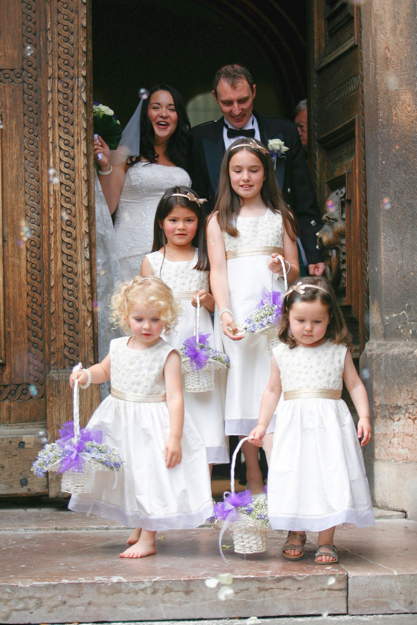 A charming blonde-haired flower girl, barefoot, scatters rose petals across the cobblestones in front of the historic town hall, leading the way for a radiant Scottish bride and groom. This picturesque scene captures the timeless beauty and tradition of their wedding procession in a setting rich with history.