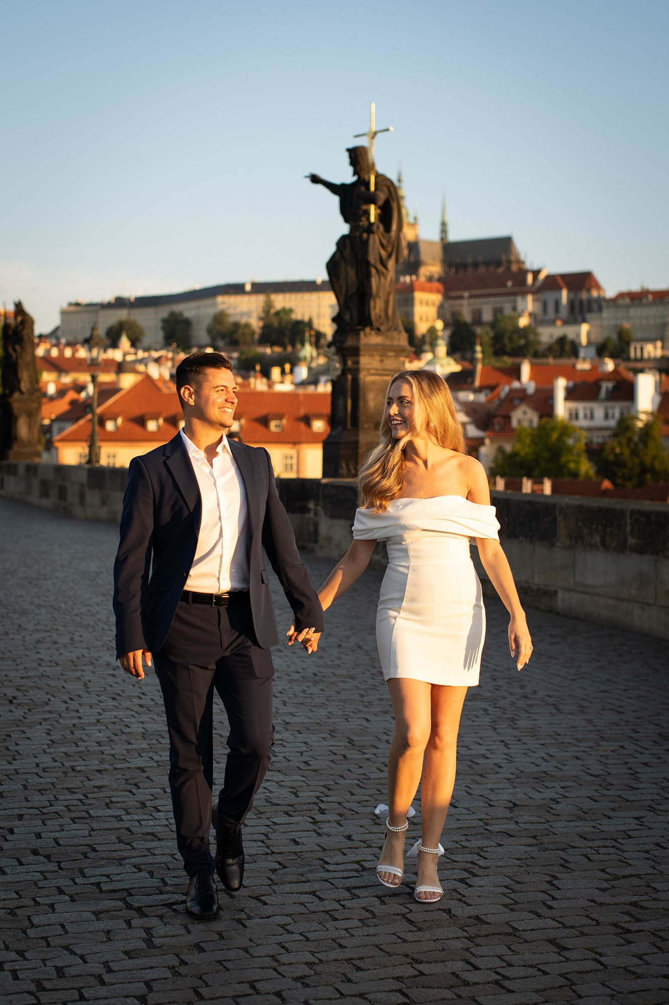 Kevin and Brooke strolling hand-in-hand on sunlit Charles Bridge Prague.