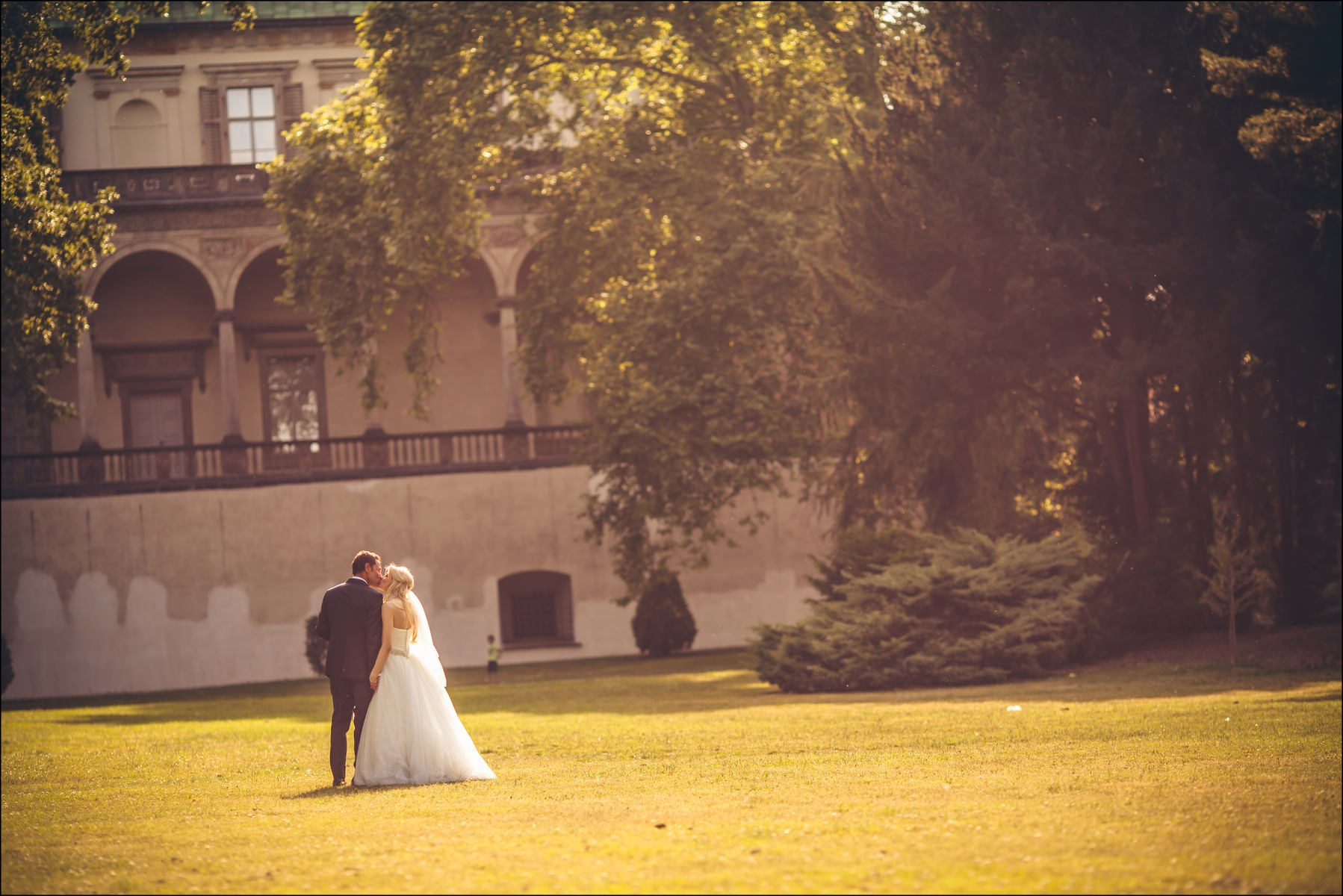 Couple walking through sun-dappled trees Prague Castle gardens wedding