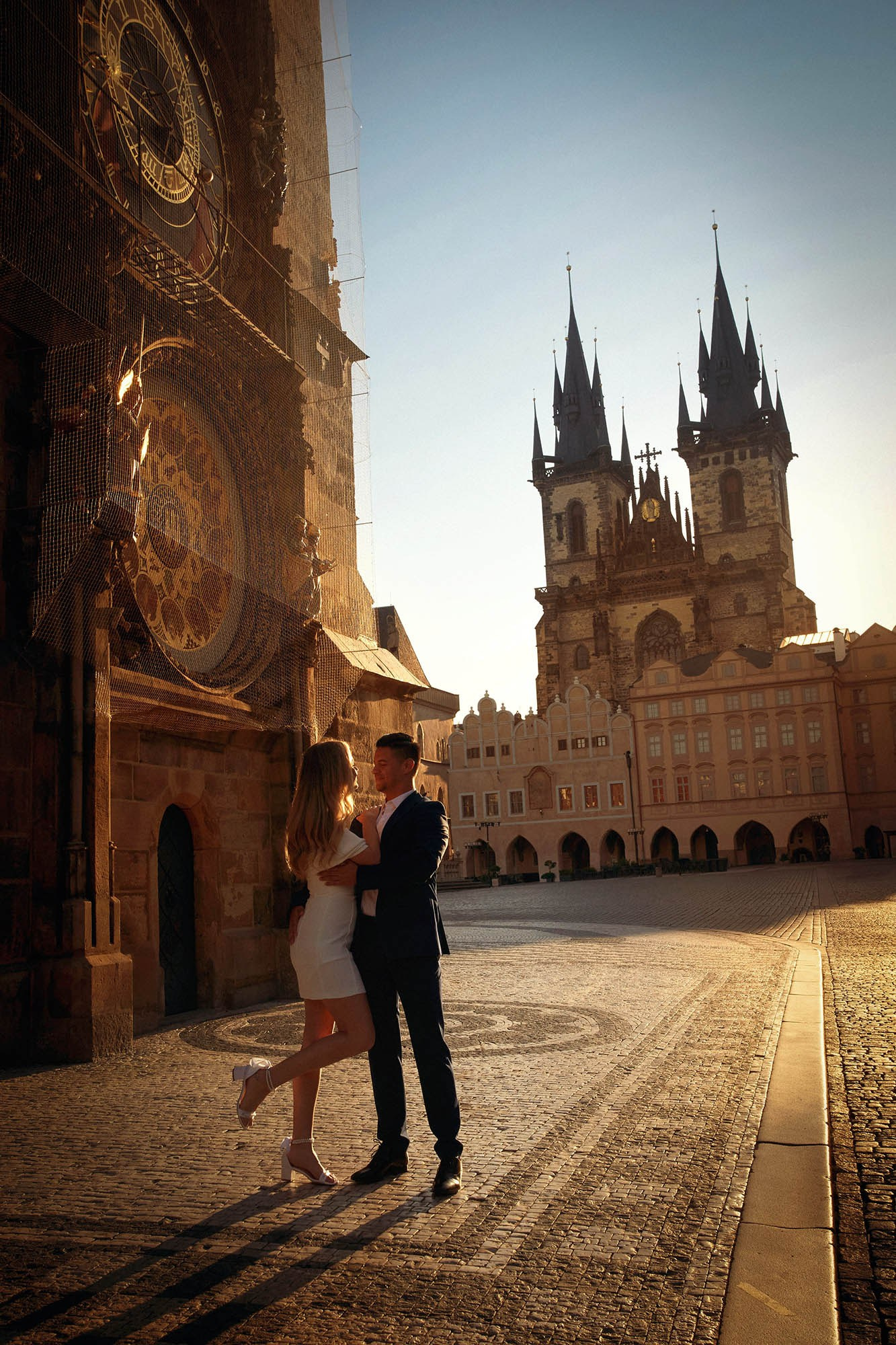 Groom lifting fiancée for a kiss under the Astronomical Clock as sun rays break near Týn Church.