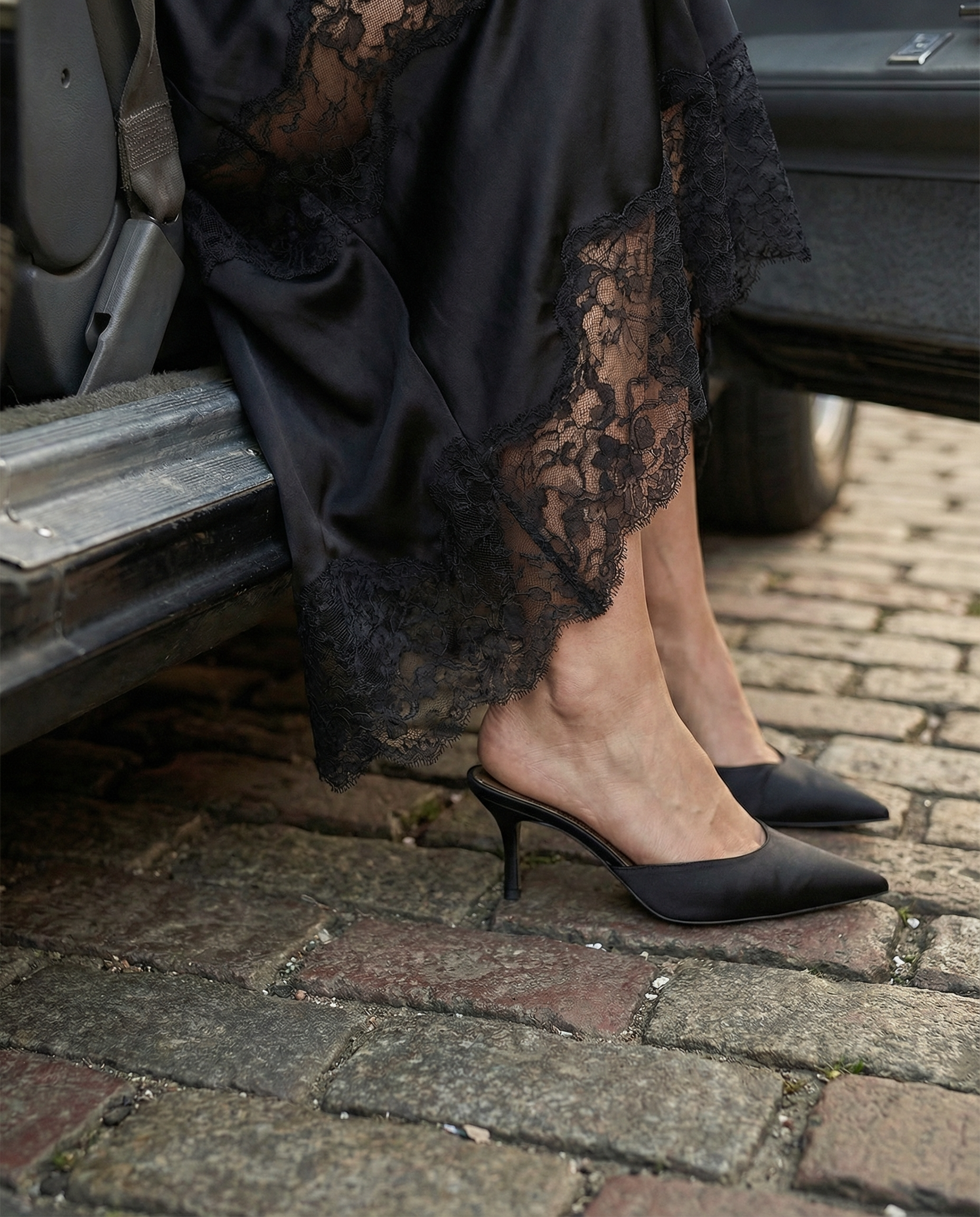 Close-up fashion detail of black satin slip dress with lace hem and black heels on cobblestone street beside a vintage car, cinematic editorial style.