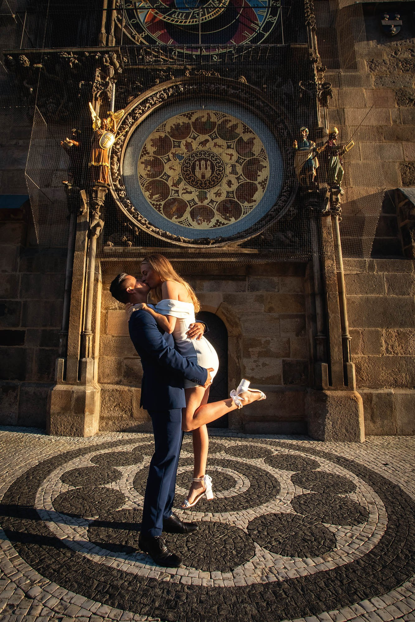 Groom lifting fiancée for a kiss under the Astronomical Clock as sun rays illuminate its historic statues and timepiece.