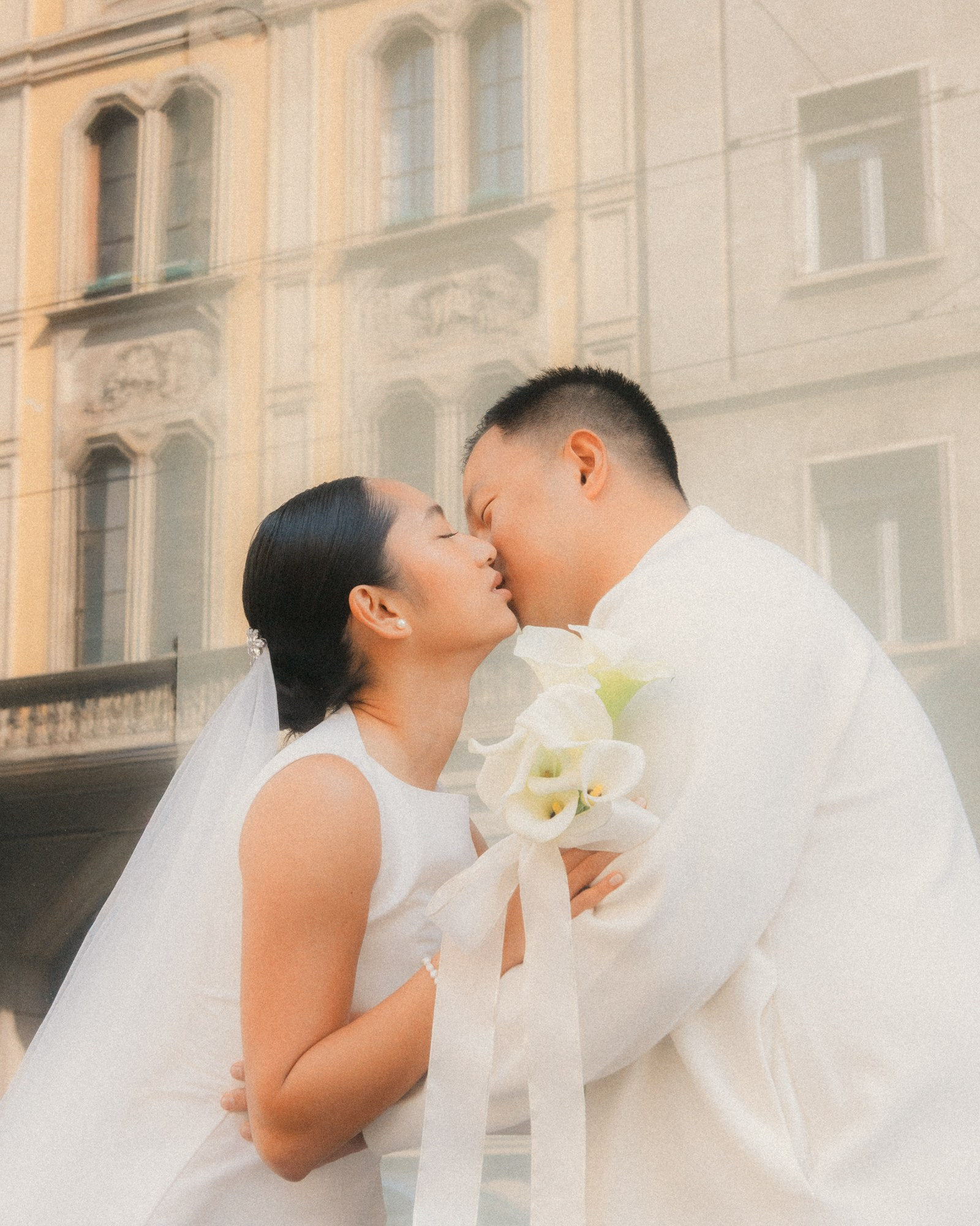Romantic wedding photo of a bride and groom sharing a tender kiss in an elegant urban setting. The bride holds a refined white calla lily bouquet, symbol of purity and timeless beauty. Emotional and natural wedding photography. wedding photographer, bride and groom kiss, calla lily bouquet, romantic wedding, wedding photography, couple portrait, elegant wedding, urban wedding, love story, destination wedding