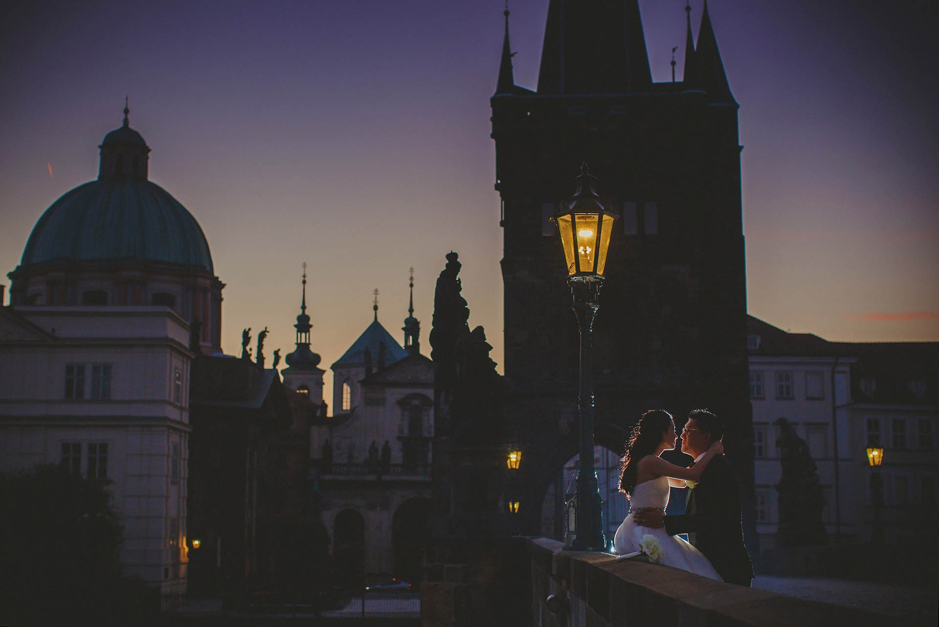 Cedric Son tuxedo embrace Catherine atop Charles Bridge dawn gas lamps Prague portrait.