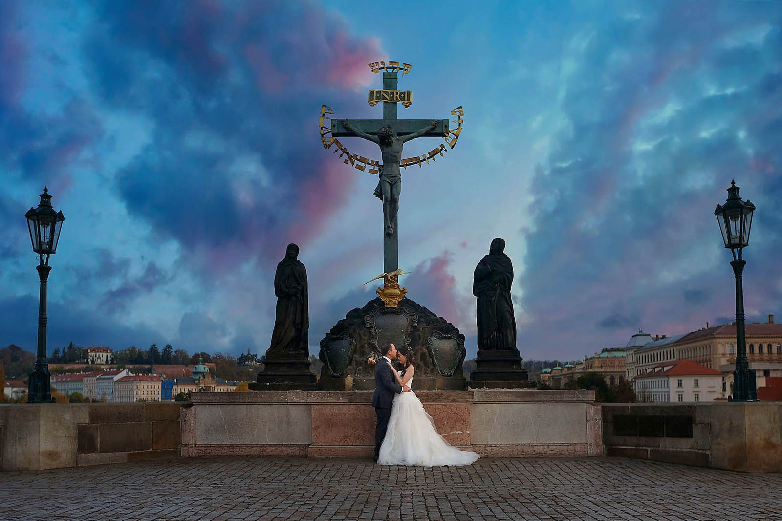 Newlyweds embrace under Charles Bridge statues as the sky flares vividly above