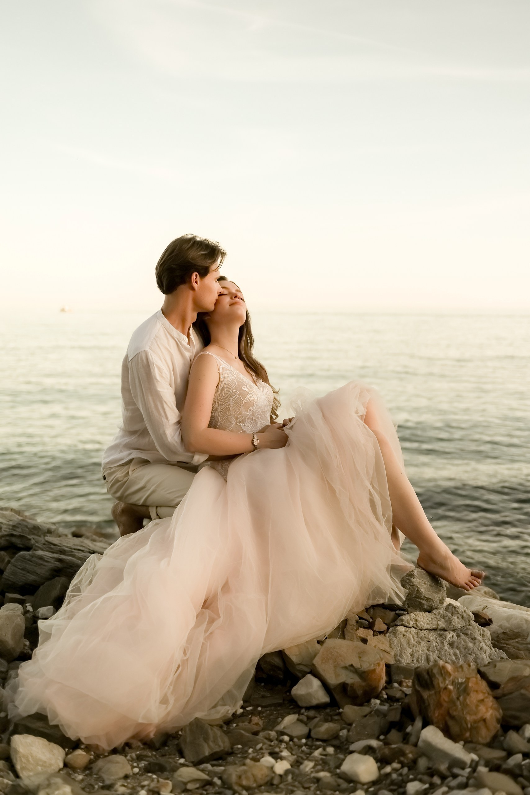 Brautpaar sitzt entspannt auf Felsen am Meer, Brautkleid im Abendhimmel, romantisches Shooting Costa Blanca
