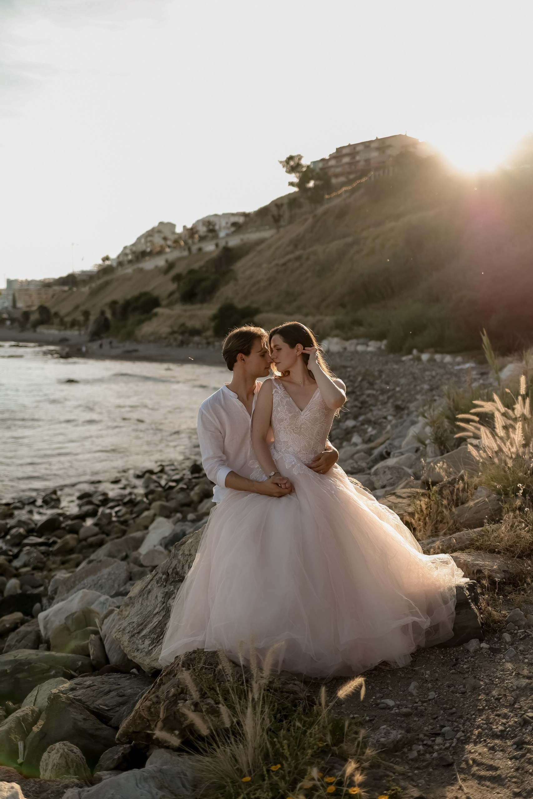 Brautpaar umarmt sich zart auf Felsen am Meer, Abendhimmel, romantische Hochzeitsfotos Costa Blanca