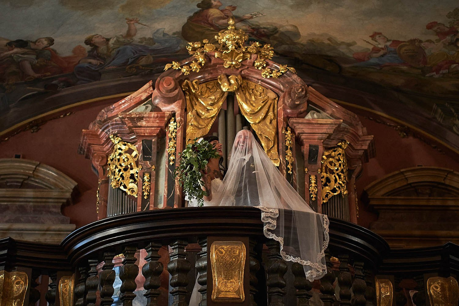Tattooed bride covering groom's face with veil on the chapel balcony.