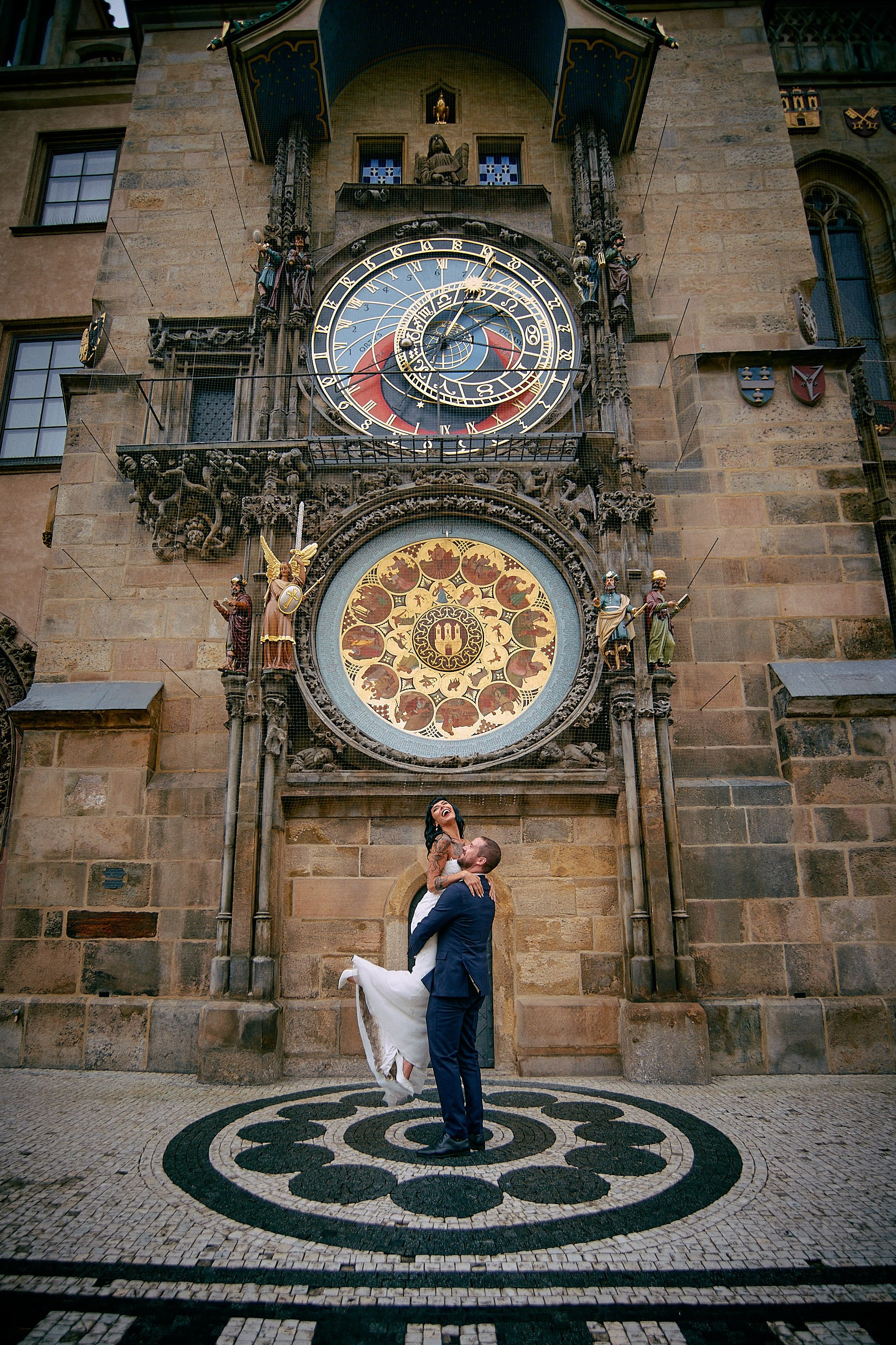 Groom lifting joyful laughing bride under medieval Prague clock.