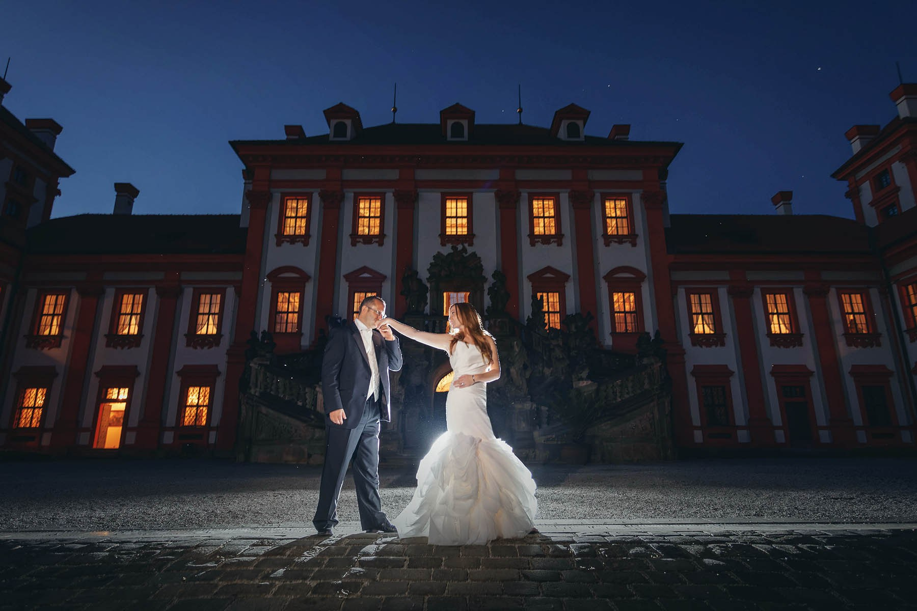A groom kisses his brides hand against the facade of the Troja Chateau during the twilight hour in Prague