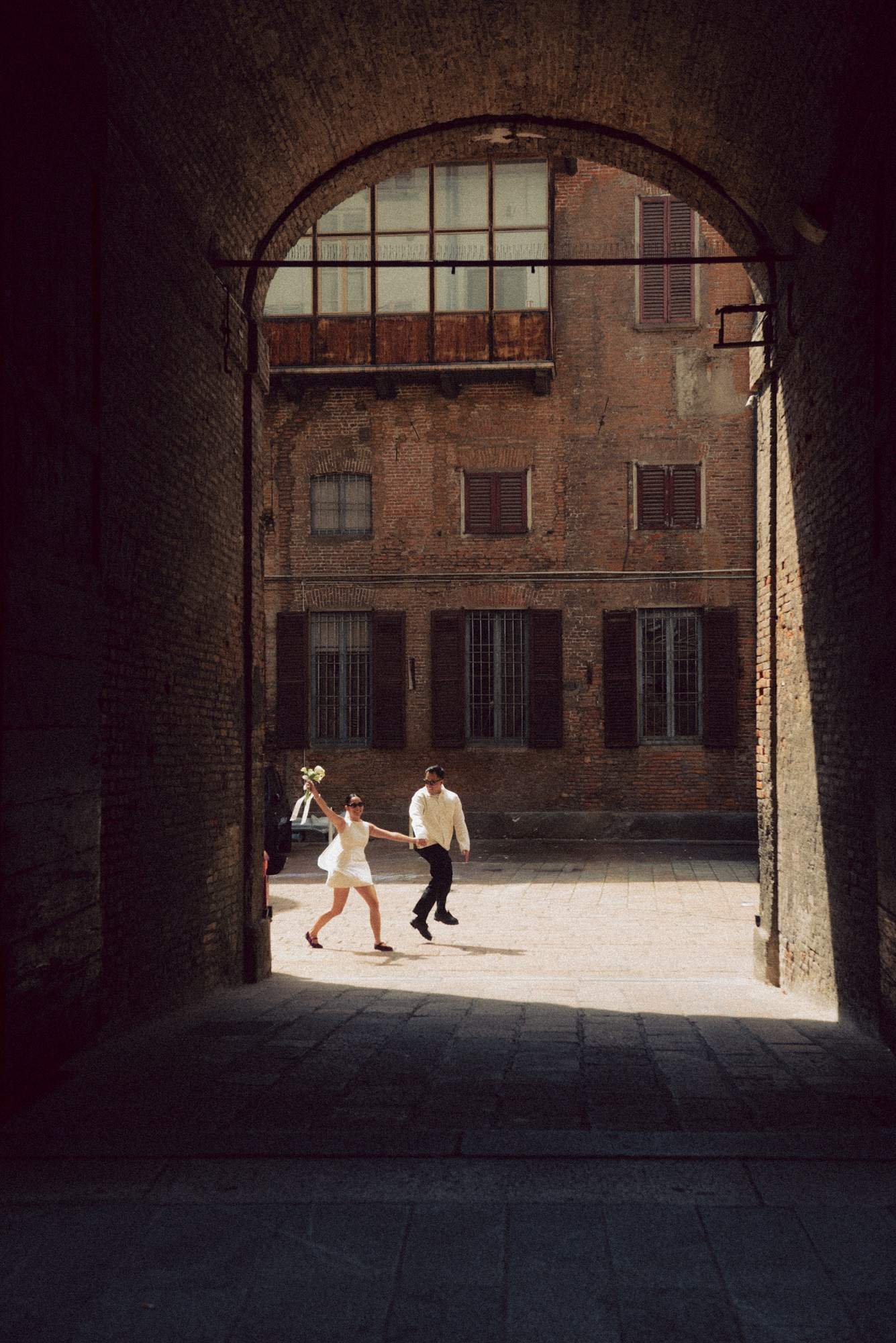 The newlyweds run joyfully hand in hand through a historic brick archway, bathed in natural light inside an Italian courtyard. A vibrant, spontaneous moment that captures freedom, love, and the beauty of modern documentary wedding photography. documentary wedding photography, bride and groom running, spontaneous wedding moment, Italian wedding, creative wedding photographer, candid wedding photo, urban wedding, love story photography