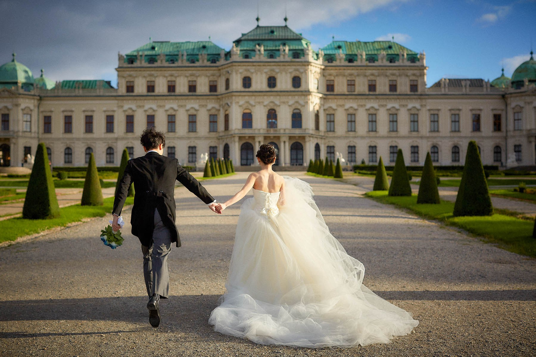 Newlyweds running hand in hand toward Belvedere Palace sunlight.