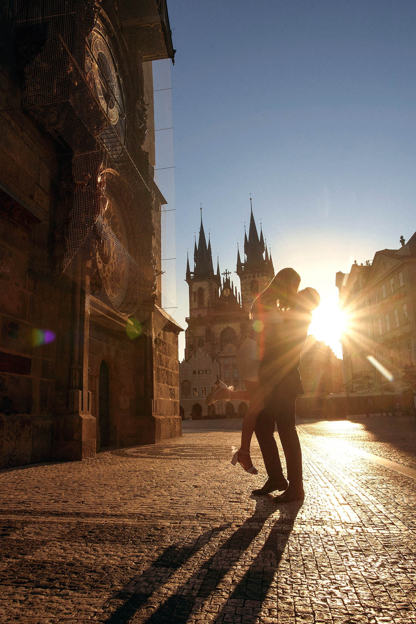 Groom lifting fiancée for a kiss under the Astronomical Clock as sun rays break near Týn Church.